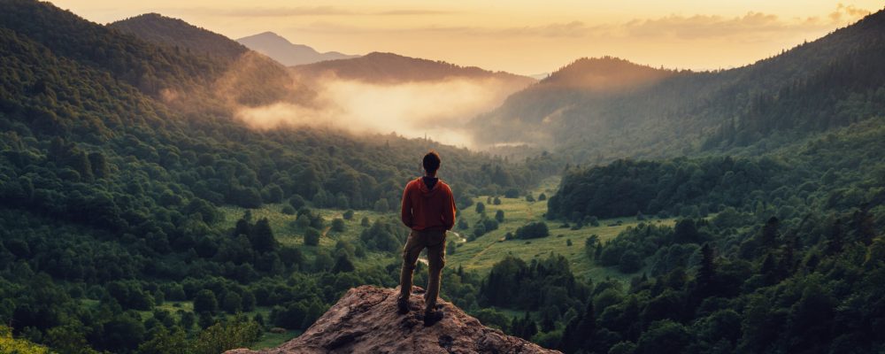 Man standing on top of cliff at sunset Young man standing on top of cliff in summer mountains at sunset and enjoying view of nature