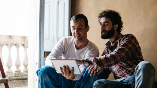 Portrait of two carpenters using tablet sitting beside opened balcony door in workshop.