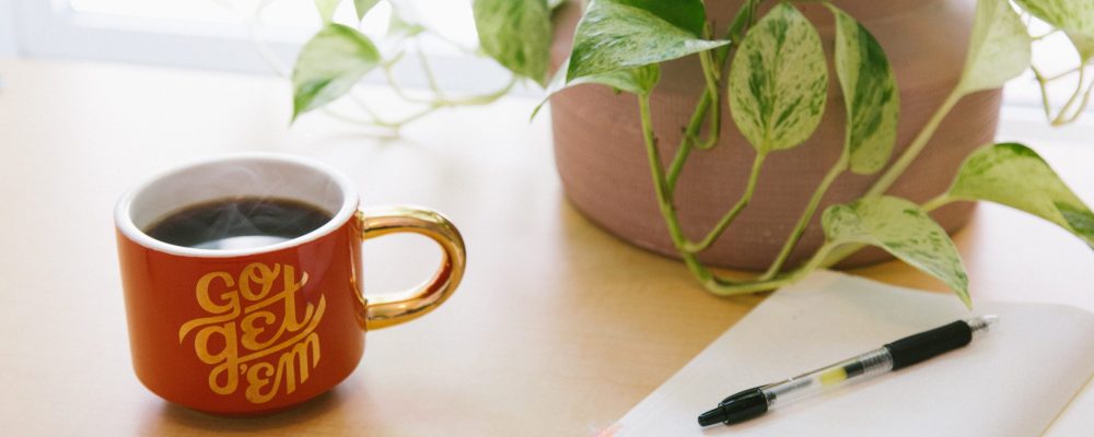 kyle-glenn-_AR74EoWdy0-unsplash Mug reading "Go Get 'Em" sits next to an open journal with a pen.