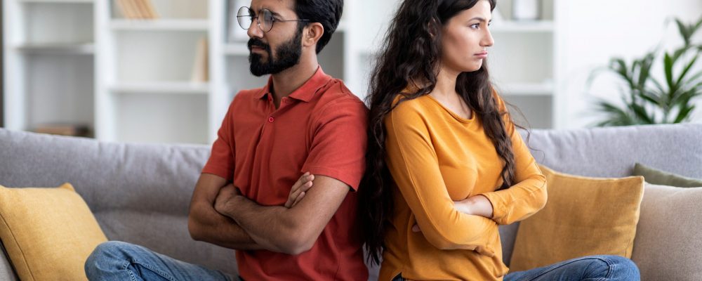 Young,Indian,Couple,Sitting,Back,To,Back,On,Couch,At Young,Indian,Couple,Sitting,Back,To,Back,On,Couch,At
